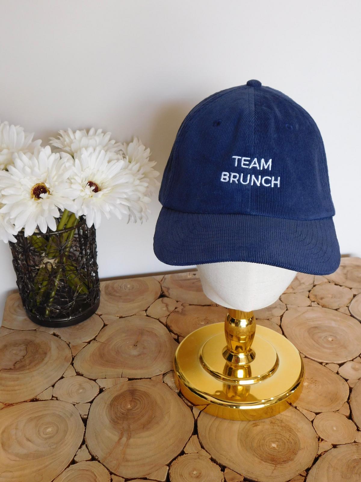 Casquette en velours côtelé bleu marine brodée Team Brunch sur une tête dorée, avec des fleurs blanches sur une table en rondins
