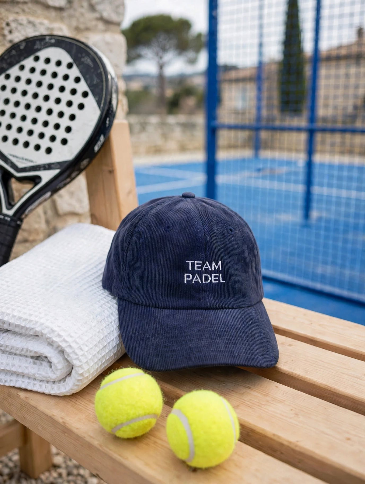 Casquette en velours côtelé bleu marine brodée Team Padel, posée sur un banc avec raquette, serviette et balles devant un terrain de padel