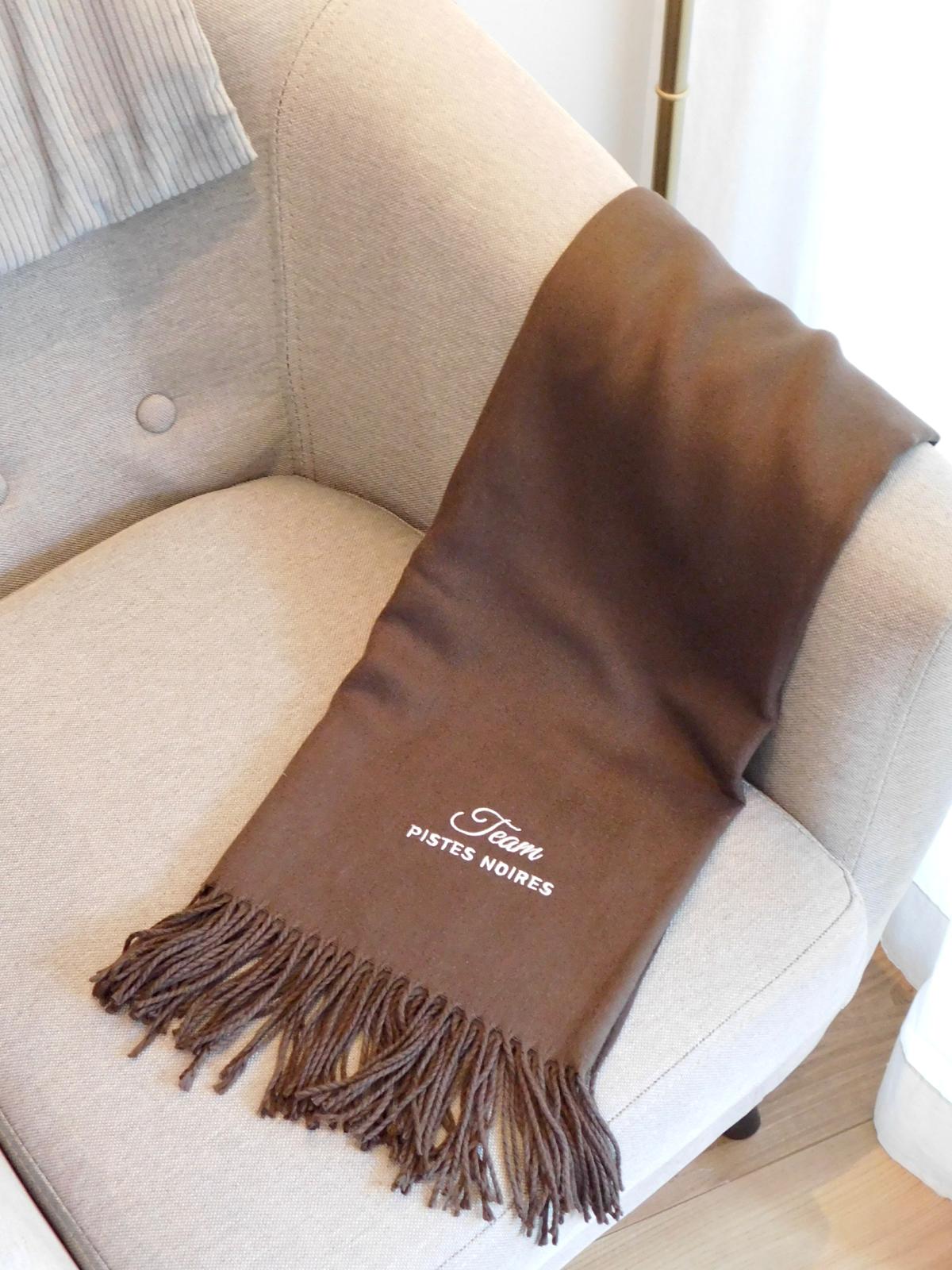 Écharpe marron à franges brodée Team pistes noires en blanc, cadeau pour fan de ski, posée sur un fauteuil