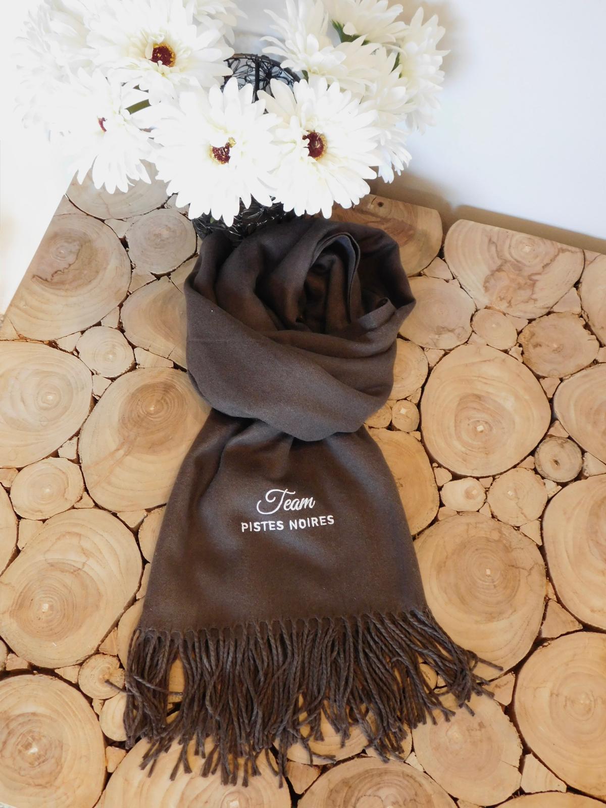 Écharpe marron brodée Team pistes noires, posée sur une table en rondins de bois avec des fleurs blanches