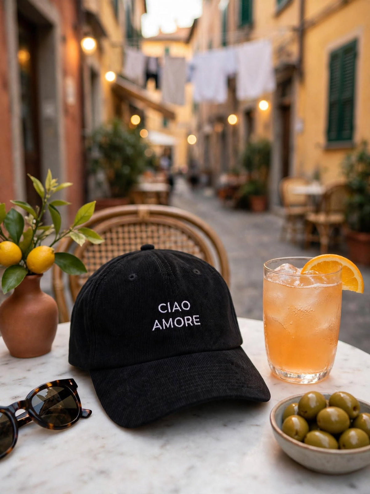 Casquette en velours côtelé noire brodée Ciao Amore sur une table de terrasse à l'italienne, avec un spritz, des olives et des lunettes de soleil
