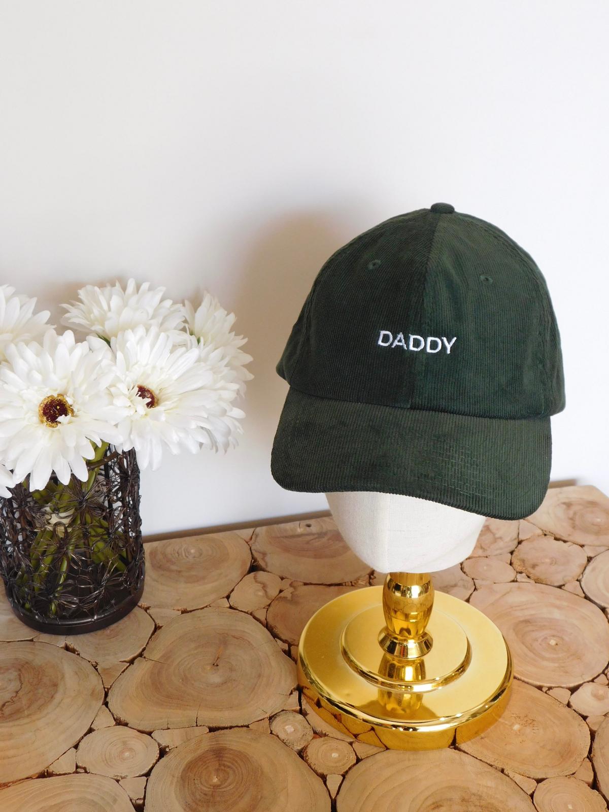 Casquette en velours côtelé vert sapin brodée Daddy sur une tête dorée, avec des fleurs blanches sur une table en rondins