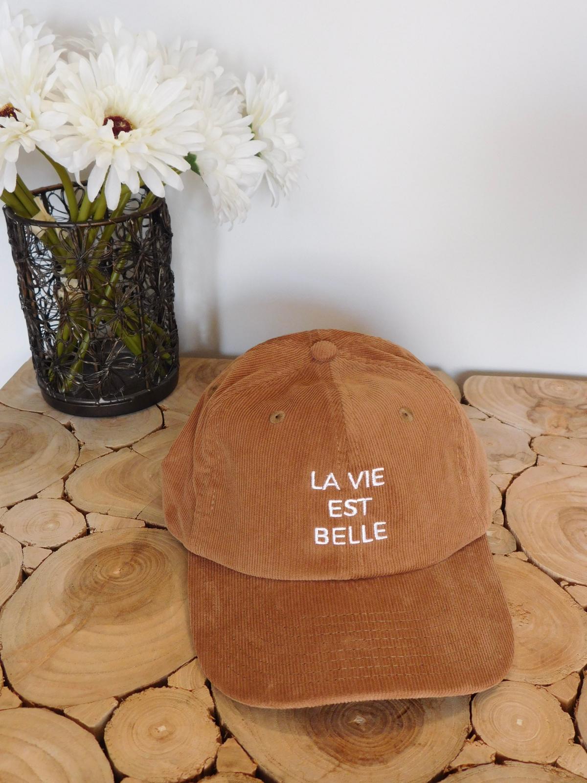 Casquette en velours côtelé camel brodée La vie est belle en fil blanc, posée sur une table en rondins avec des fleurs blanches