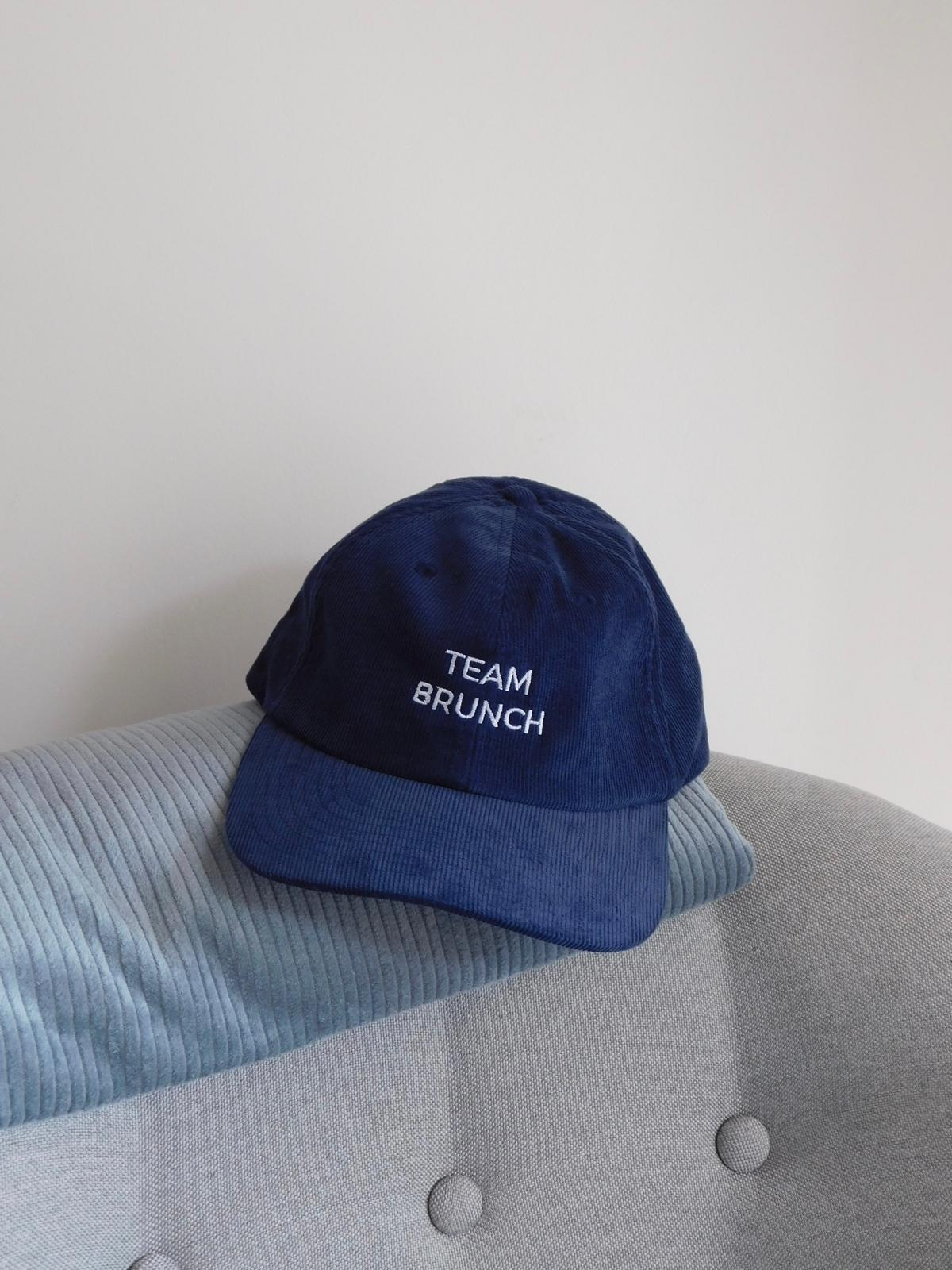 Casquette en velours côtelé bleu marine brodée Team Brunch en fil blanc, posée sur un fauteuil gris