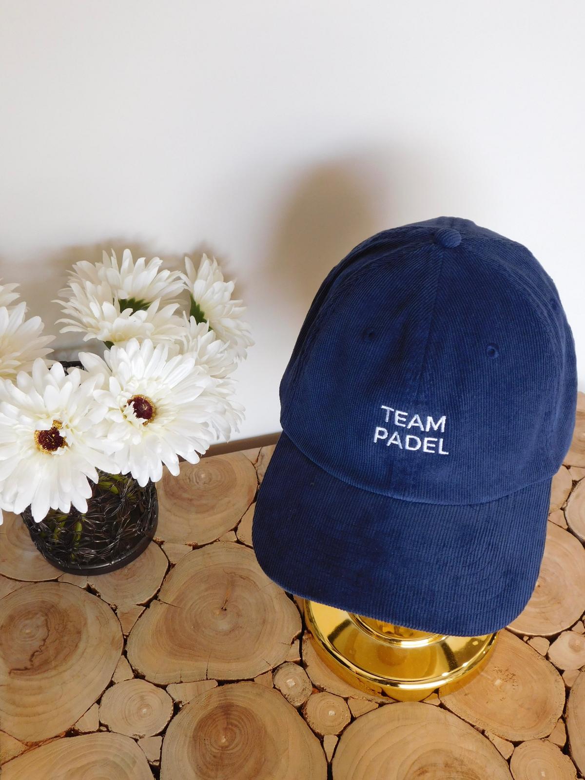 Casquette en velours côtelé bleu marine brodée Team Padel sur une tête dorée, avec des fleurs blanches sur une table en rondins